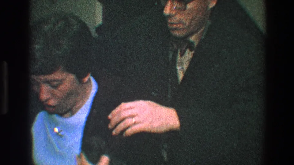 The woman in the light blue dress looks down as a man stands beside her, holding the coat in this Toledo 1968 home-movie frame.
