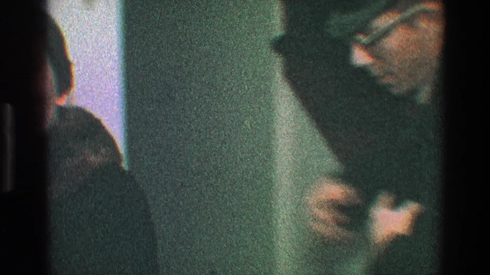 A man leans forward in the dim room as the coat-helping action continues, with figures partly cropped by the 8mm frame.