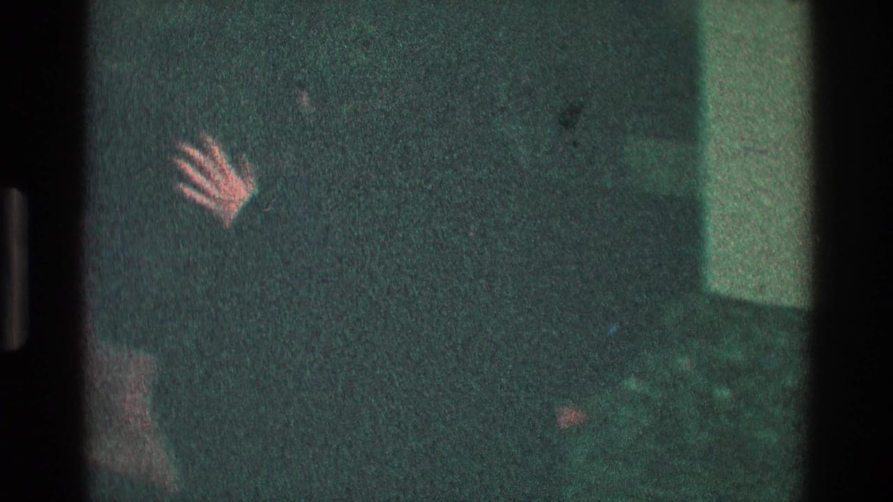 Adult in dark clothing raises a hand in a dim room near a wall.
