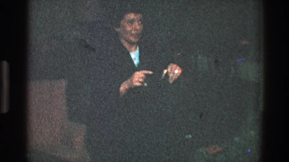 In the 1968 Toledo interior, an adult wearing a dark coat and hat gestures with both hands near a sofa or chair.