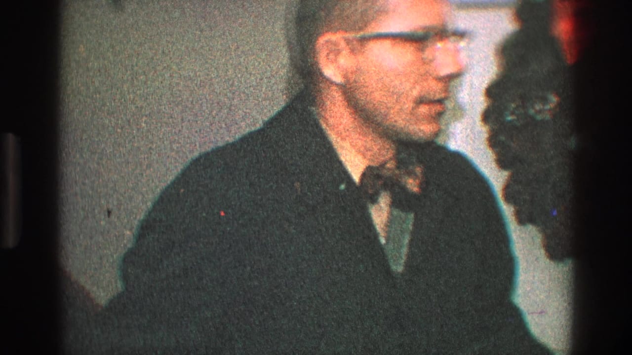 In 1968 Toledo home-movie footage, a man in glasses and a dark jacket stands near a wreath with a red bow on a white door.