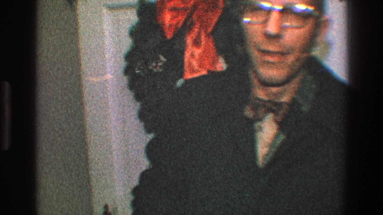 Man facing the camera beside a dark wreath with a red bow on a white door.