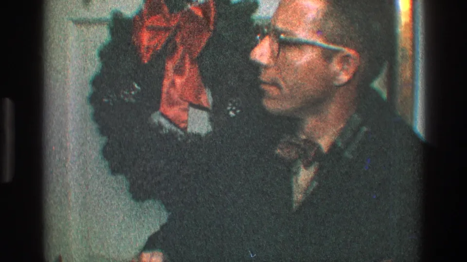 A side view shows the man in glasses beside the Christmas wreath and red bow on a white paneled door.