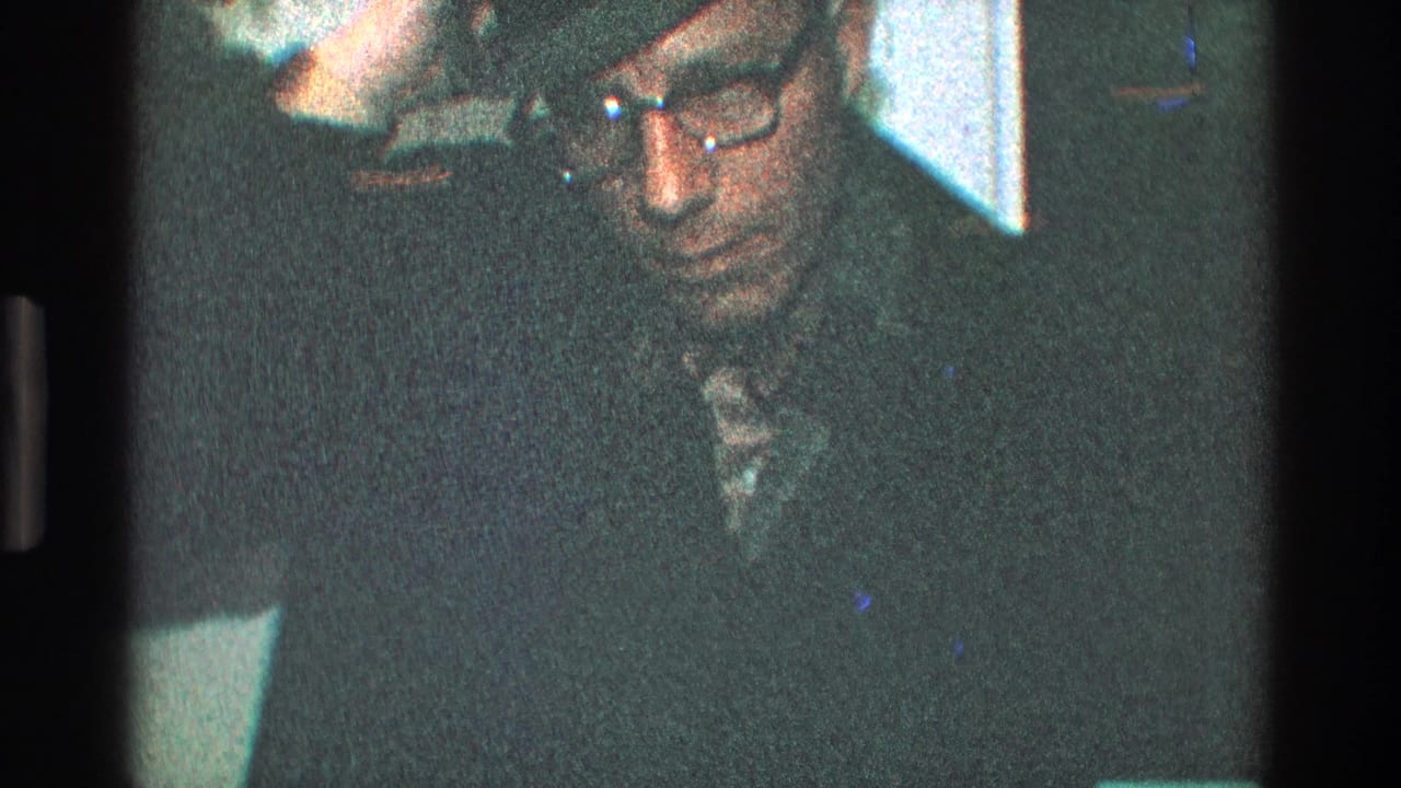 Man in a dark hat and glasses looks downward in a dim interior.