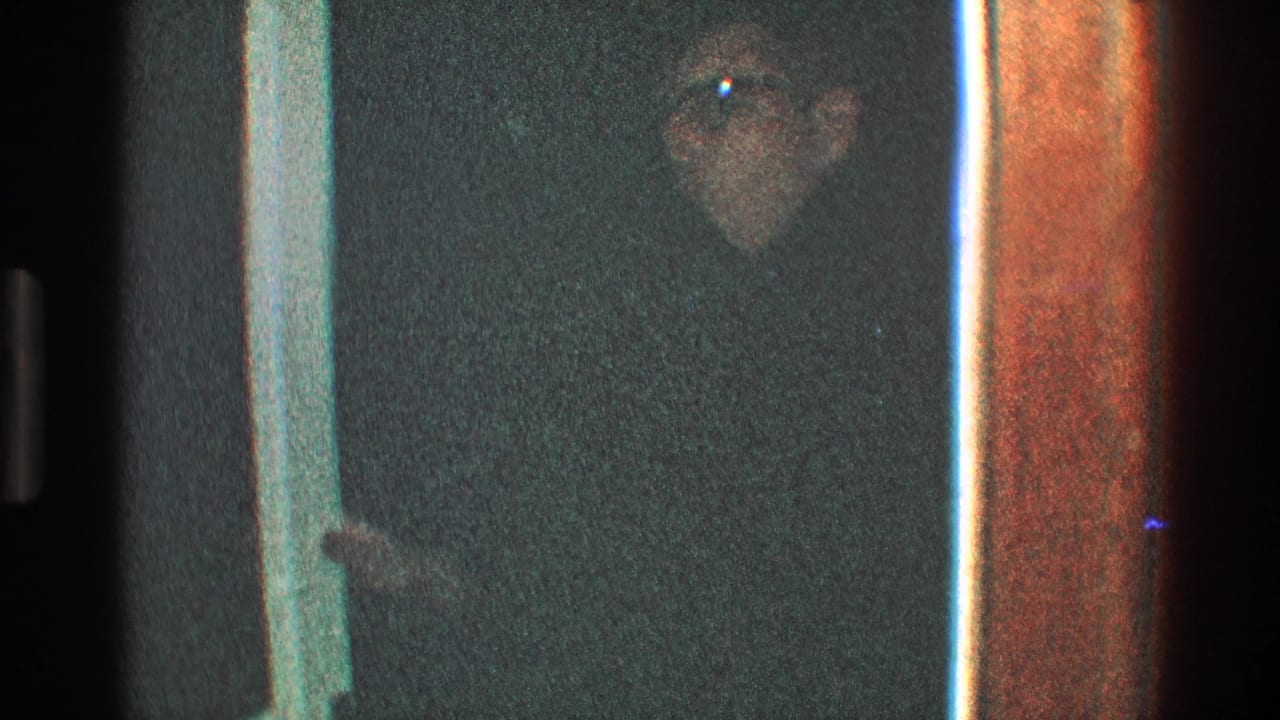 Man in glasses and a dark coat stands partly shadowed near a doorway.