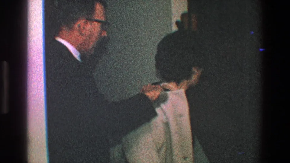 A man in a dark coat and glasses stands inside a Toledo home near another adult with dark hair and a light-colored garment.