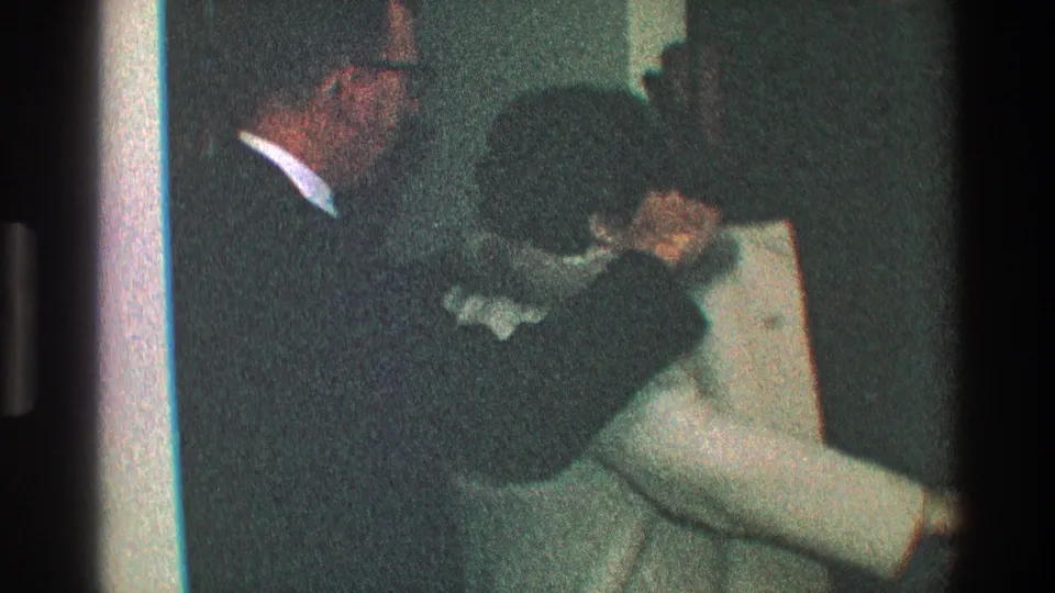 In the 1968 Toledo home interior, the dark-coated man leans close to another adult near the doorway, possibly during a goodbye.