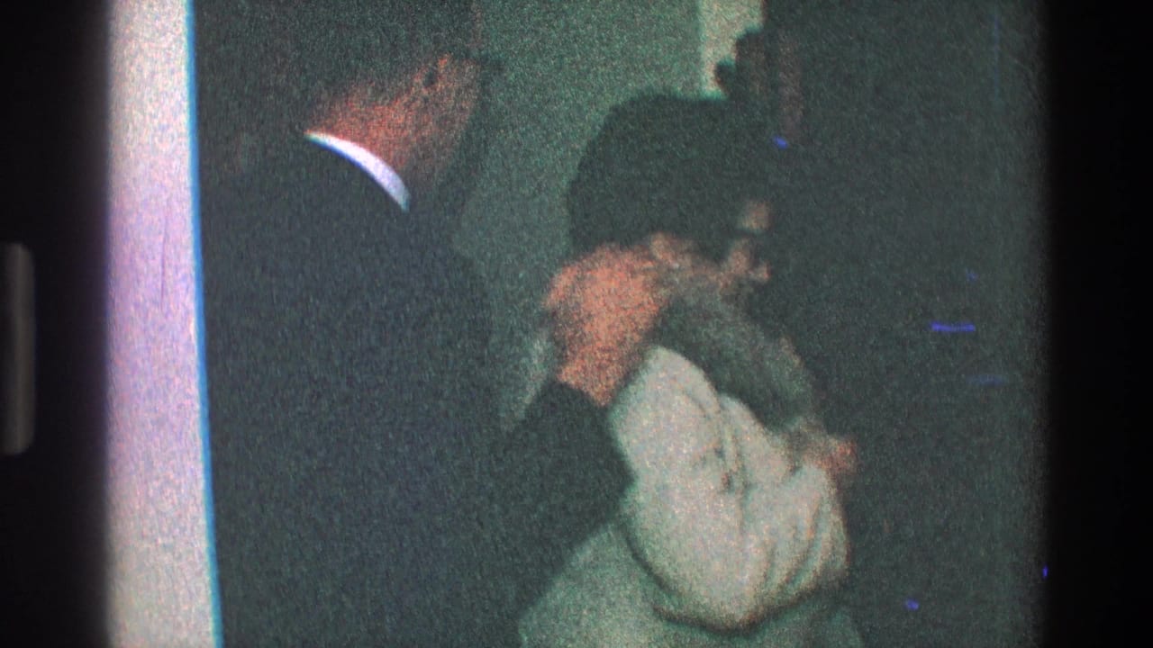 Woman in a light coat adjusts a fur hood near a doorway as a suited man stands beside her.