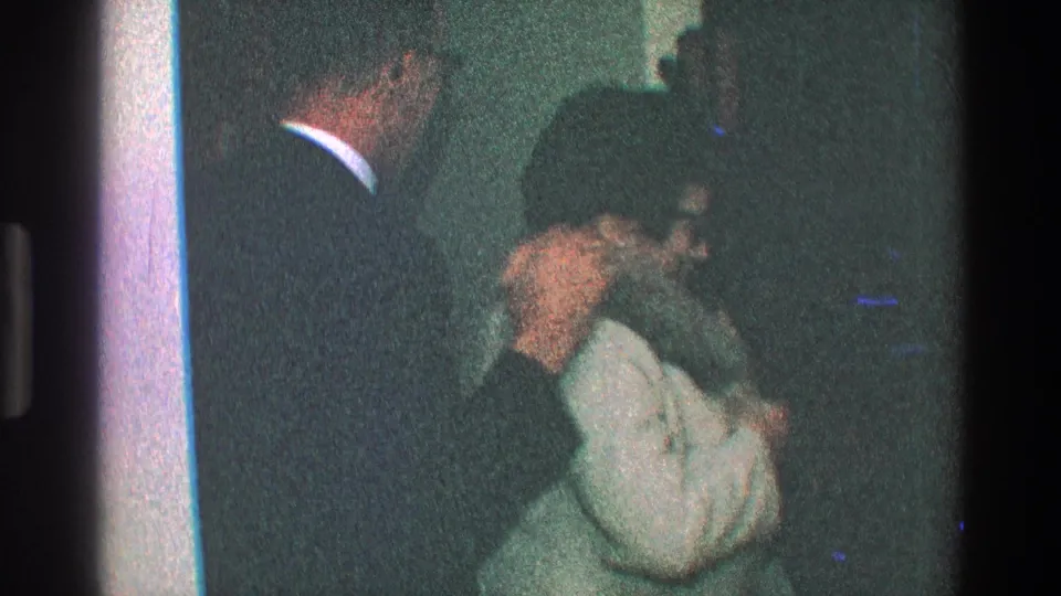 In 1968 Toledo home-movie footage, a woman in glasses adjusts a fur-trimmed hood near a doorway beside a partially visible suited man.