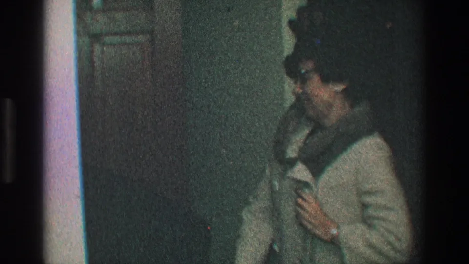 A woman in a light fur-trimmed coat and glasses looks down while adjusting the front of her coat in a private interior doorway.