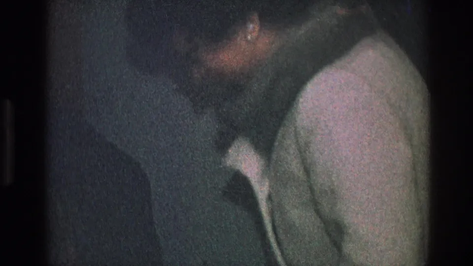 A close, grainy 8mm frame shows the man beside the woman in a light coat during the indoor Toledo scene.
