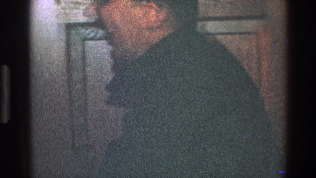 Man in glasses and dark suit stands in profile near a paneled door.