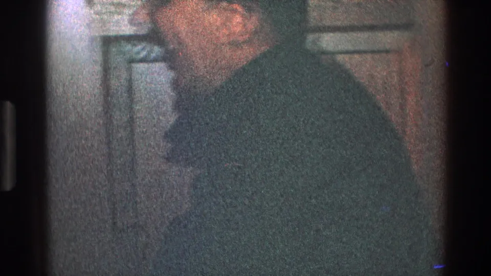 A man in glasses and a dark suit stands in profile beside a paneled door in 1968 Toledo 8mm footage.
