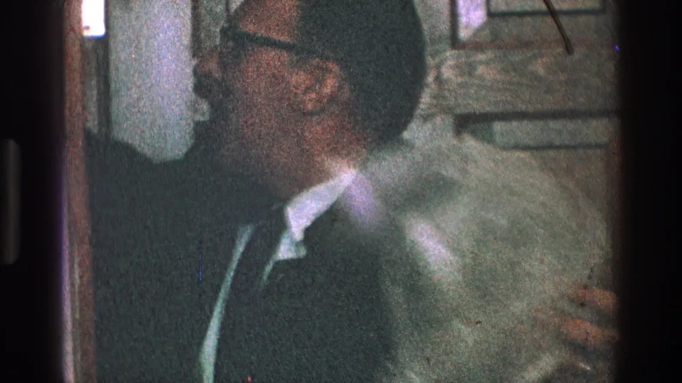 A suited man in glasses reaches near the doorway as a pale garment or indistinct object appears at the right edge of the 1968 Toledo frame.