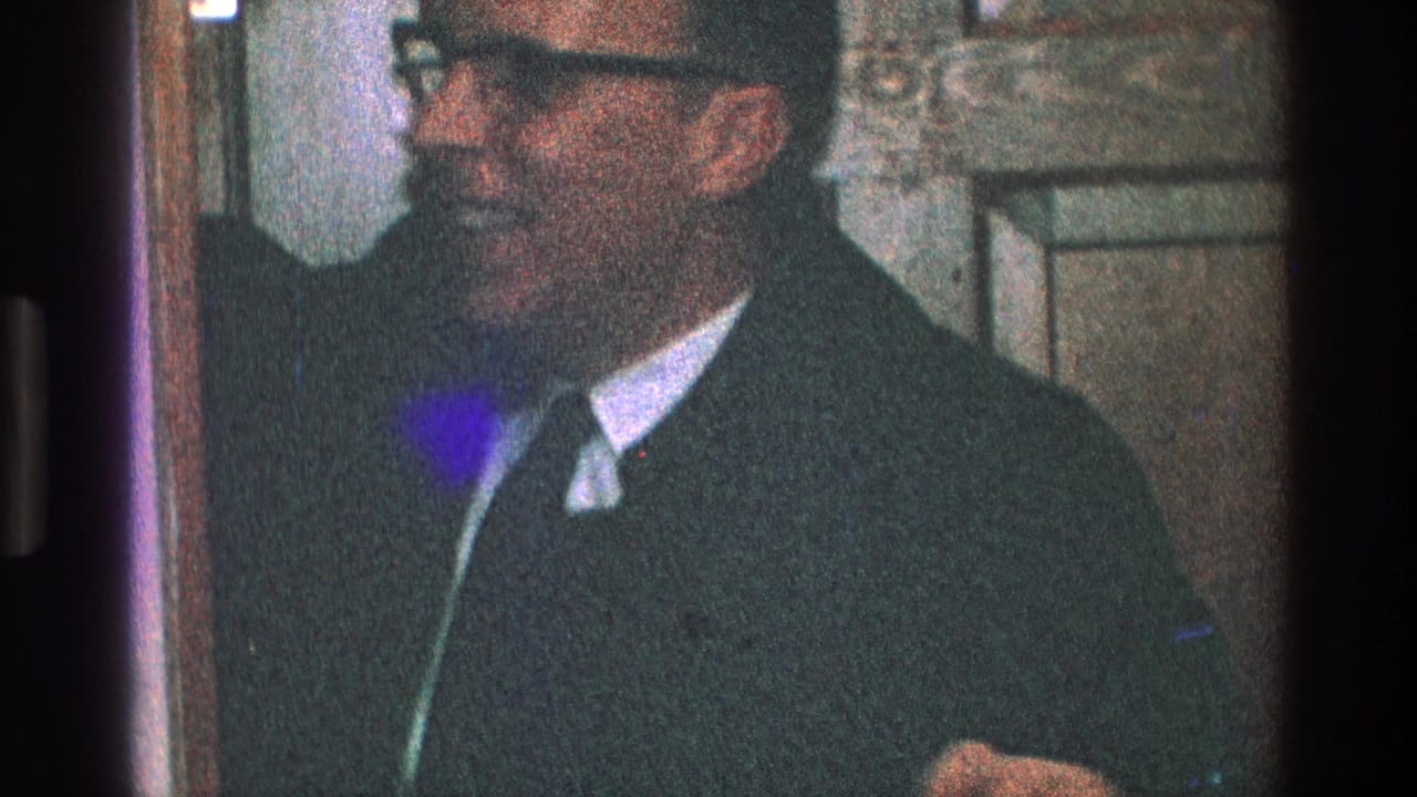 In 1968 Toledo home-movie footage, a man in glasses and a dark suit smiles near a wood interior door.