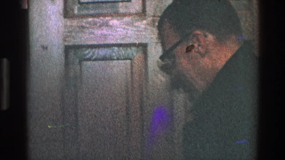 The suited man turns toward the wood door or closet, his glasses and profile partly visible in the grainy 8mm frame.