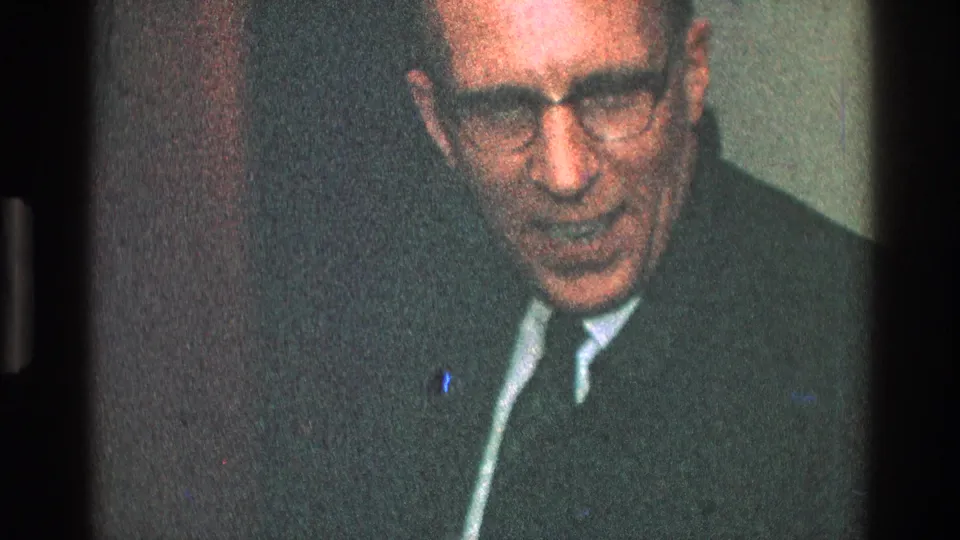 The man in glasses looks downward with a focused expression, wearing a dark suit and tie in the Toledo interior scene.