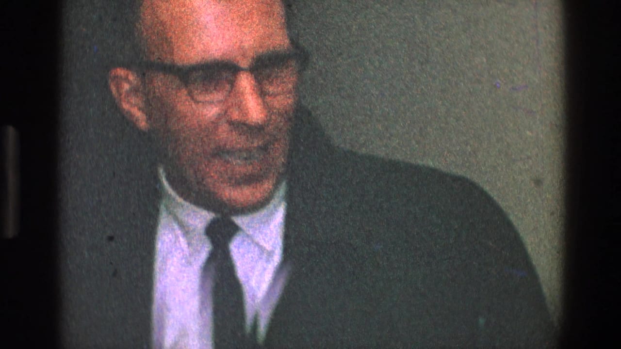 Older man with glasses in a dark jacket and tie speaks indoors.