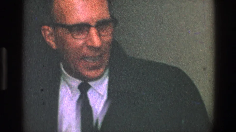Older man in glasses, dark jacket, and tie appears to speak indoors in 1968 Toledo home-movie footage.