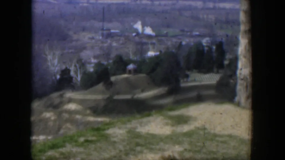 1951 Texas 8mm frame looking over a sloped landscape with evergreens, bare trees, open ground, and distant buildings.