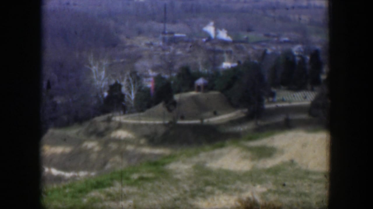 1951 Texas 8mm frame from a hilltop showing open ground, a road, trees, scattered houses, and hazy terrain beyond.