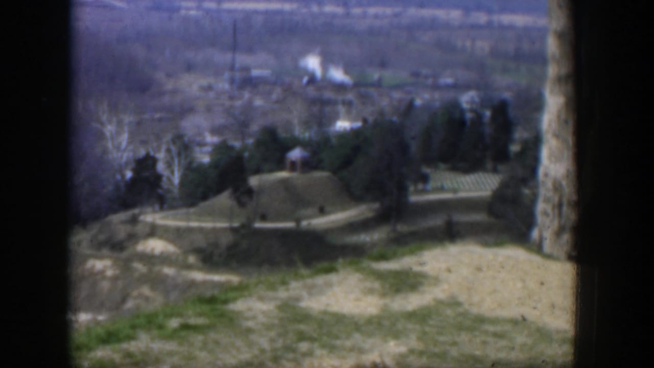 Hilltop landscape with a road, small houses, trees, open ground, and distant haze.