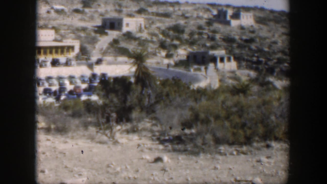 1951 Texas 8mm frame of a hillside property with low buildings, rows of parked cars, scrub vegetation, and an access road.