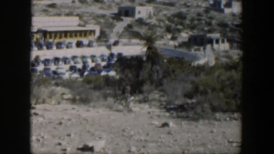 1951 Texas 8mm frame showing a parking area of mid-century cars beside a long low building on a dry hillside.