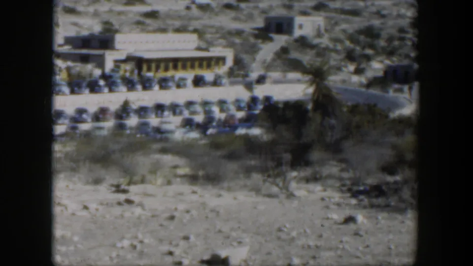 1951 Texas 8mm frame with parked cars, a low roadside building, hillside terrain, brush, and open foreground ground.