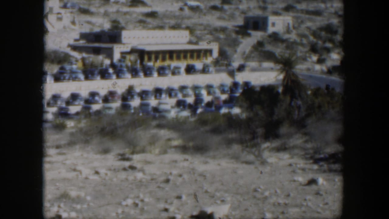 1951 Texas 8mm frame showing rows of classic cars parked by a low building in a dry roadside landscape.