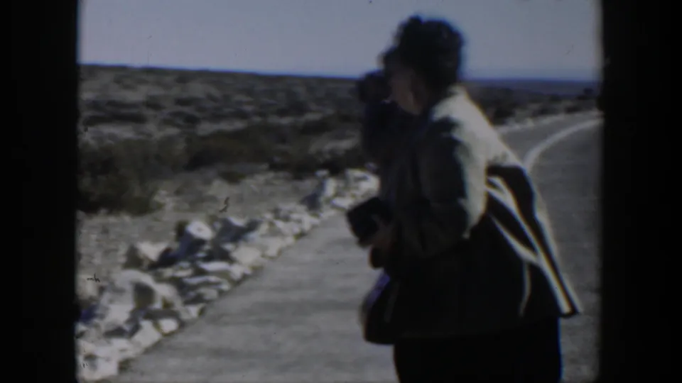 In the Texas desert roadside scene, a traveler stands near a paved road with scrubby hills and sky behind.