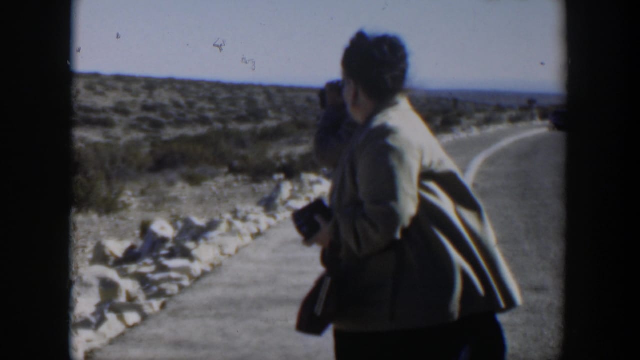 Woman holding a camera stands on a desert road with another person partly behind her.