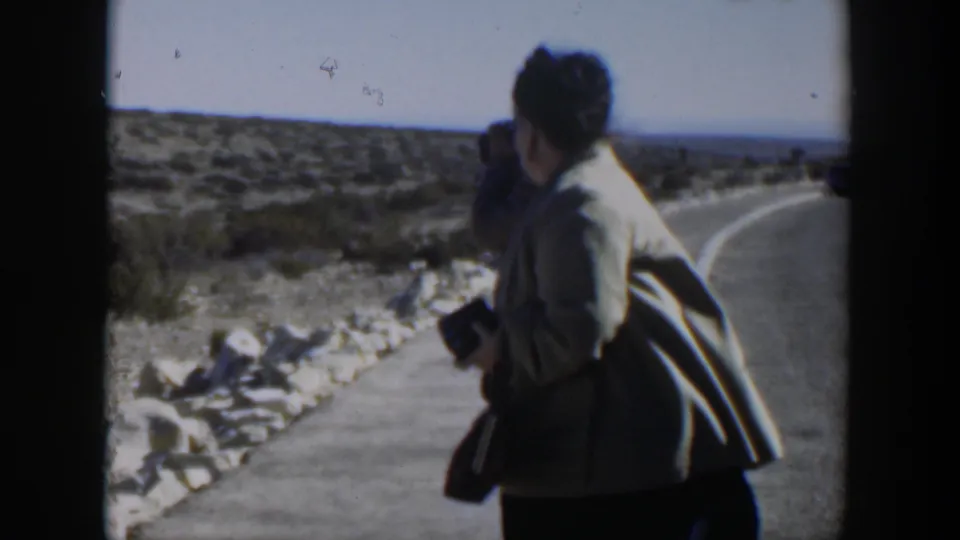 In Texas 1951 home-movie footage, a woman in a light jacket holds a camera beside a curving desert road as another adult stands partly obscured.