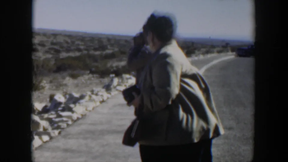 The woman remains near the Texas desert road with a camera in hand, partly blocking another adult against low shrubs and a distant horizon.