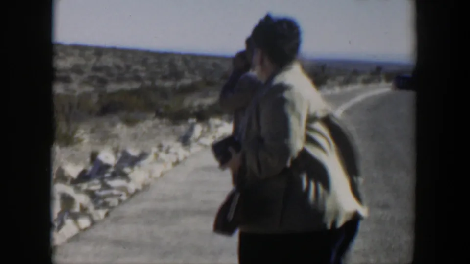 A woman in a light jacket stands with her camera on a Texas desert roadside, with another adult close behind and scrubland beyond the pavement.