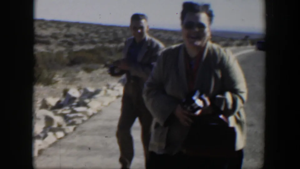 In the clearest frame, a smiling woman faces the movie camera while holding a still camera, with another adult and camera behind her on a Texas desert road.