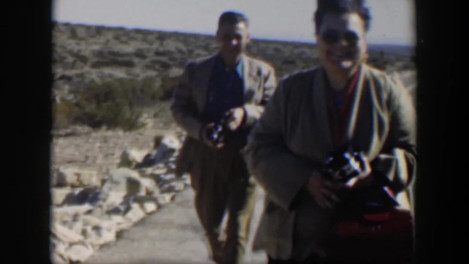 In 1951 Texas home-movie footage, two adults carrying cameras approach the viewer near a desert roadside or visitor area.