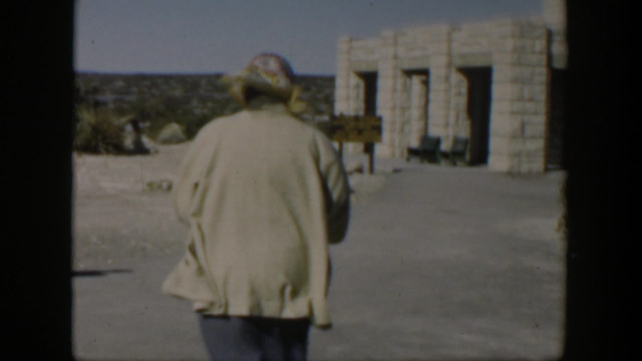 A woman in a light coat and patterned headscarf walks away from the camera toward a stone building in the Texas desert setting.