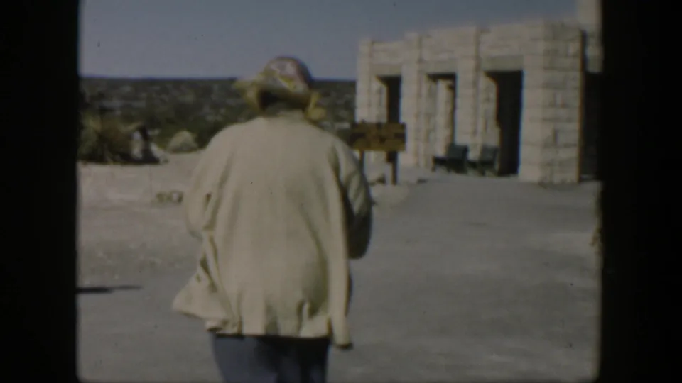 A woman in a light coat and patterned headscarf walks away from the camera toward a stone building in the Texas desert setting.
