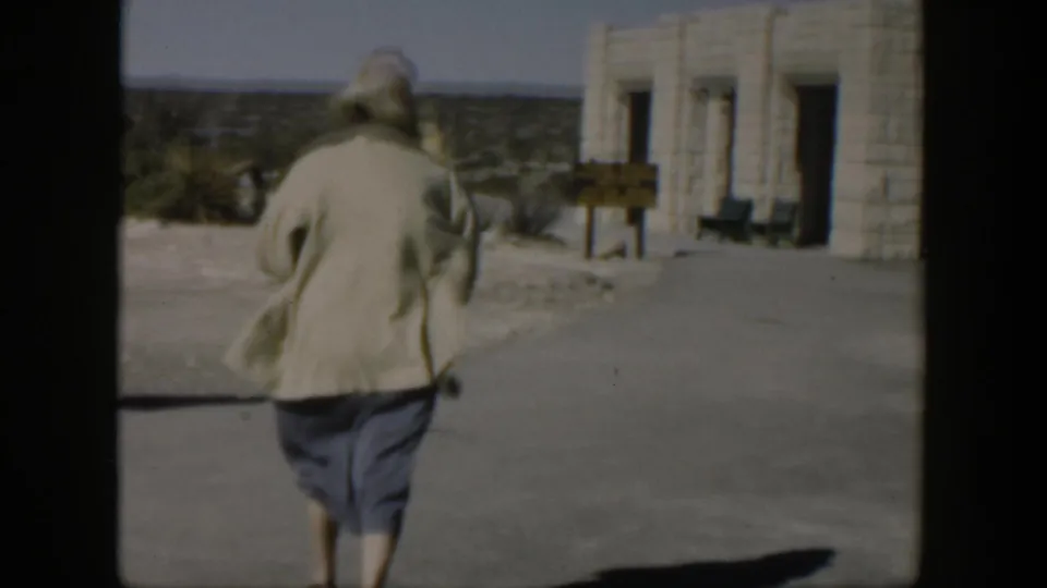The woman continues across the paved area toward the stone building, with open desert terrain visible beyond it.
