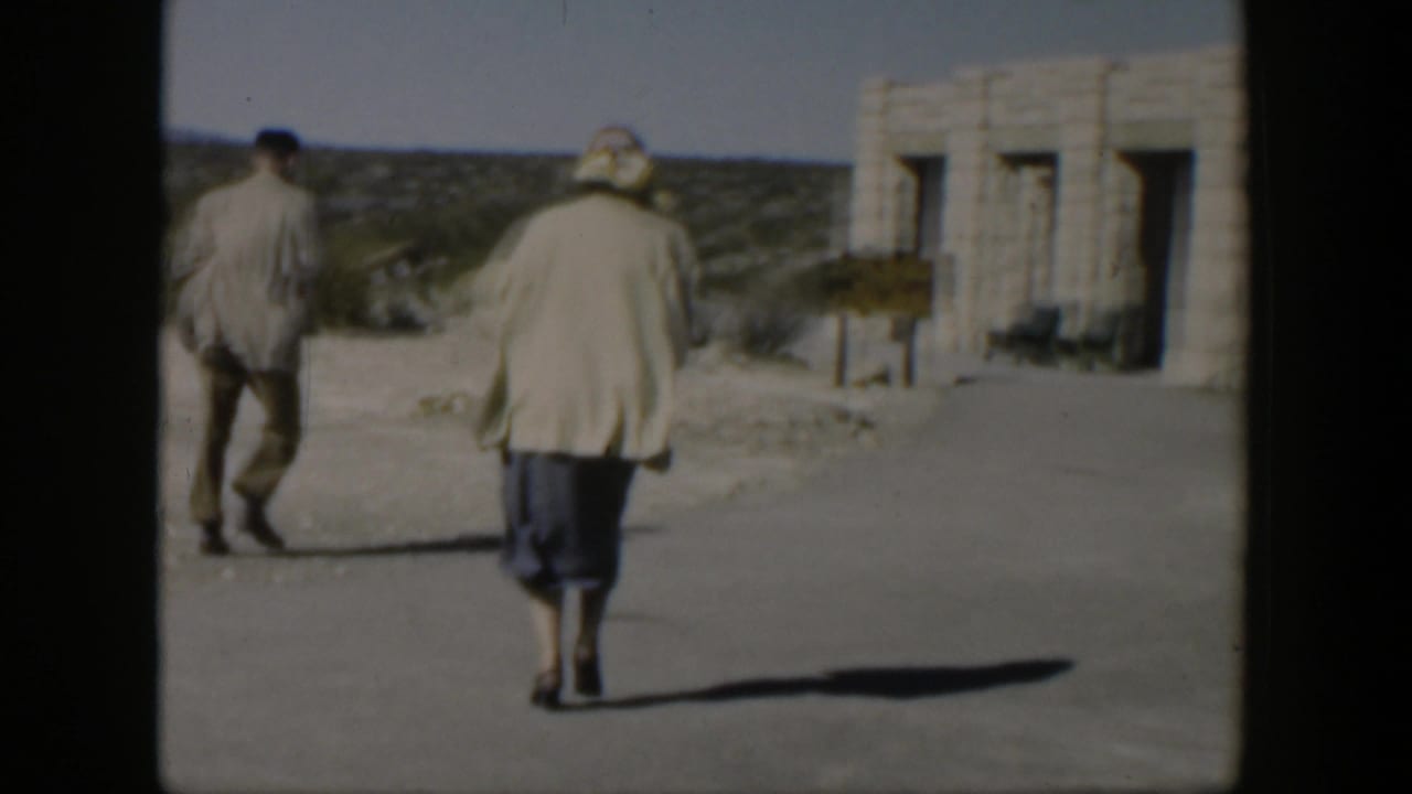 Two adults walk away from the camera toward a pale stone building in bright sun.