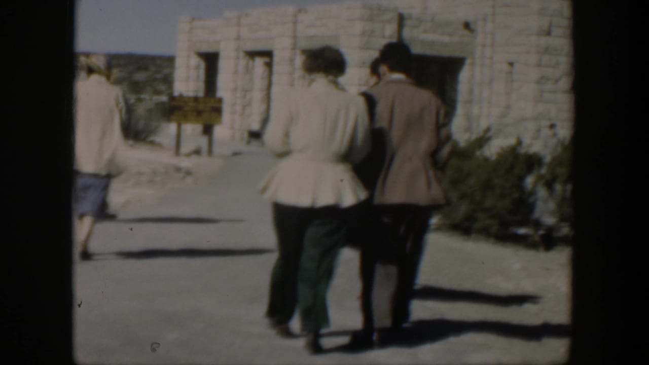 Several adults walk near a pale stone building with arched doorways.
