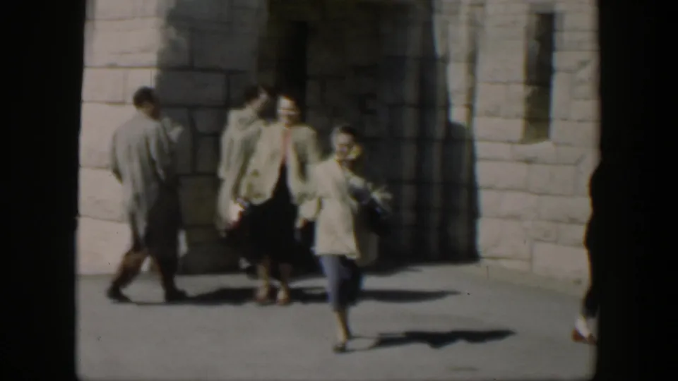 1951 Texas 8mm frame showing adults moving around a pale stone facade as a woman walks toward the camera.