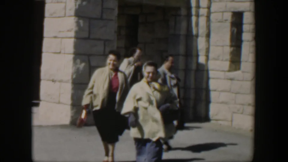 In 1951 Texas home-movie footage, two women in midcentury clothing walk past a white stone building with deep doorway shadows.