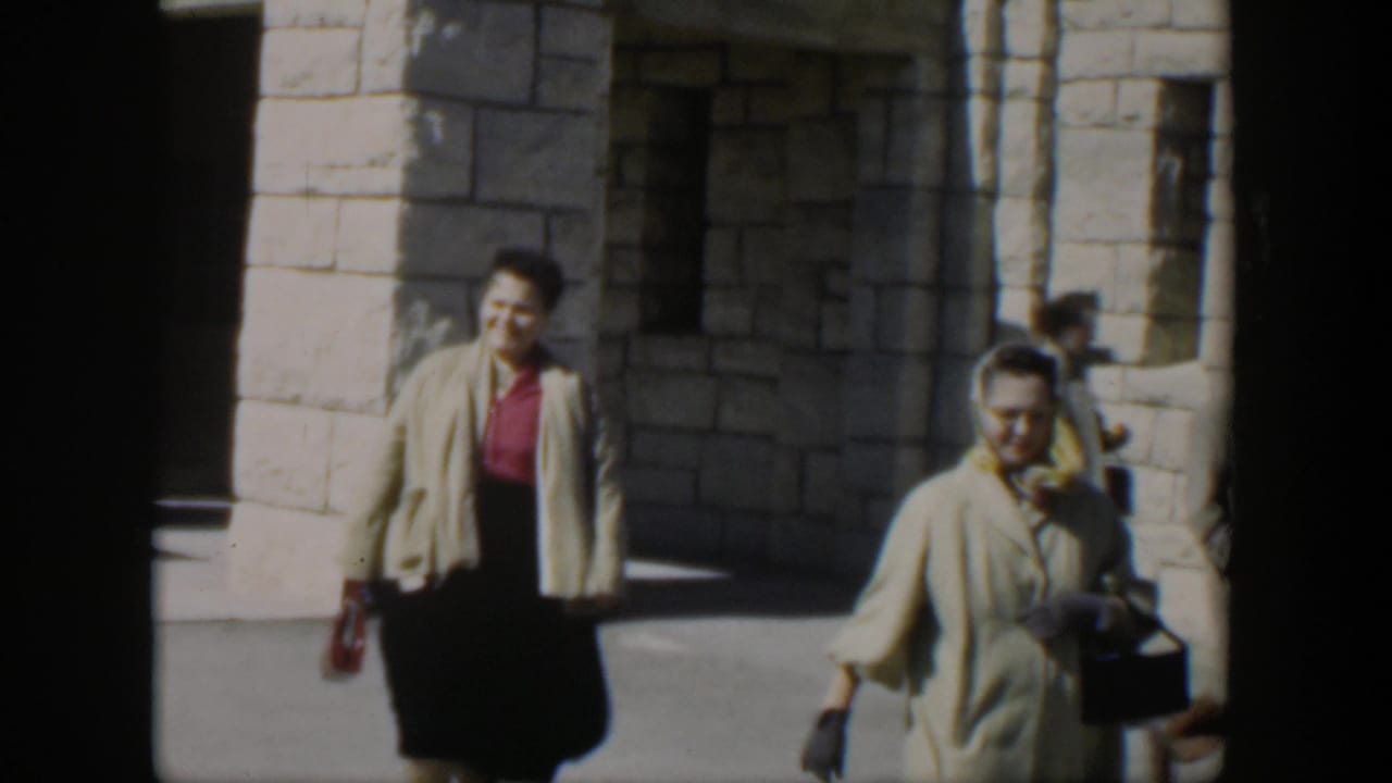 A woman in a beige cardigan walks beside another woman near stone arches.