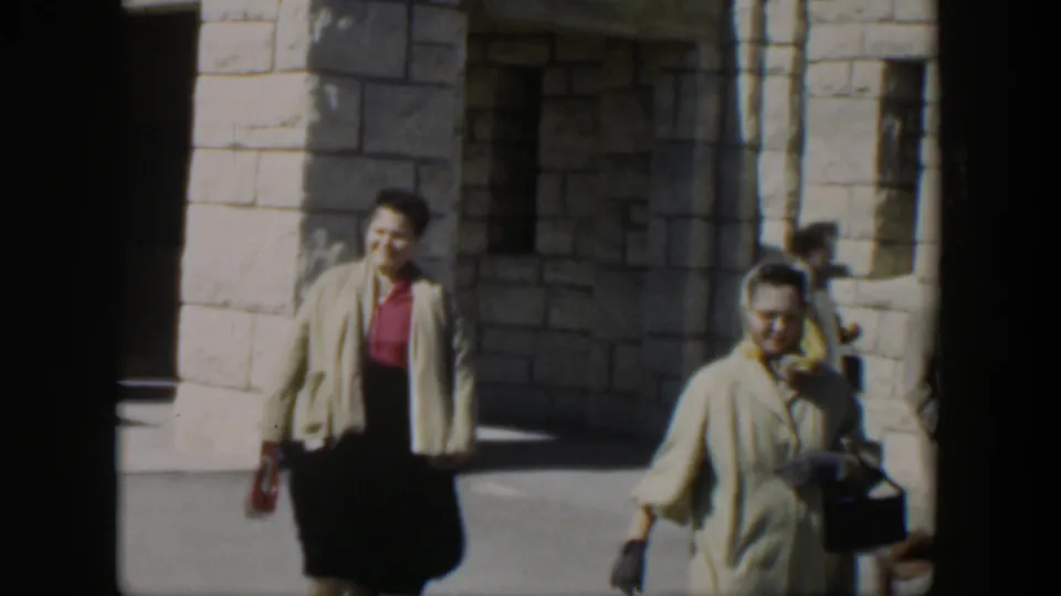 A woman in a beige cardigan, red top, and dark skirt walks in sunlight near a white stone building in Texas archival footage.