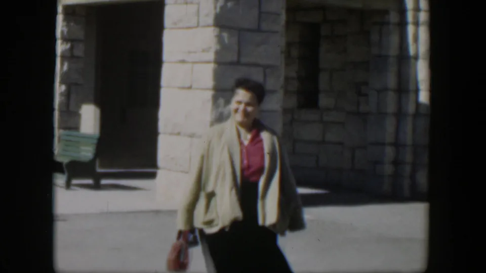 The frame shows a woman in 1950s dress carrying a small handbag near a white stone wall and shaded doorway in Texas.