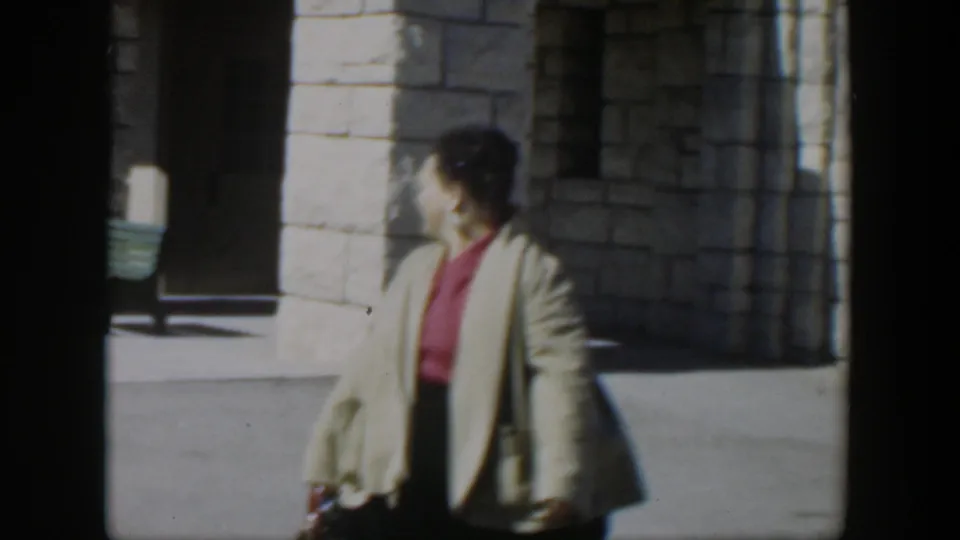 A woman in a beige cardigan and red top turns in profile while walking past the sunlit stone building in 1951 Texas footage.
