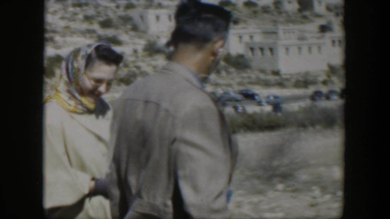 Woman in a headscarf stands beside a man on a sunny roadside with hillside buildings behind them.