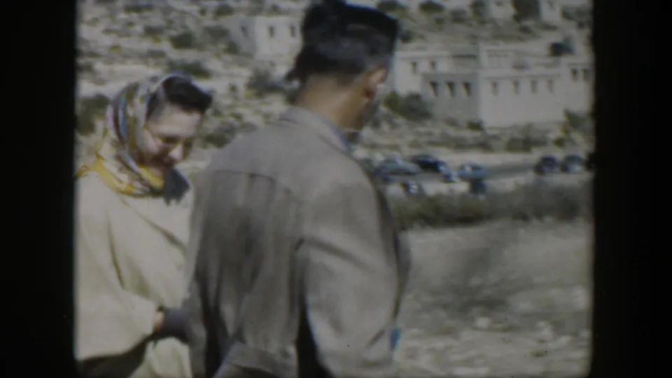 In 1951 Texas home-movie footage, a woman in a headscarf stands near a man on a roadside below a dry hillside with buildings and cars.